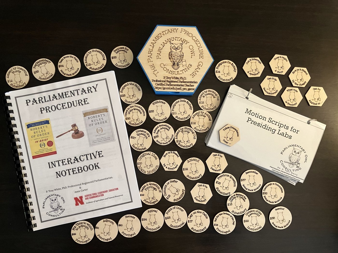 Laser-cut wooden motion chips, hexagonal prompt tiles, a printed Motion Scripts deck, and the Parliamentary Procedure Interactive Notebook laid out on a table.